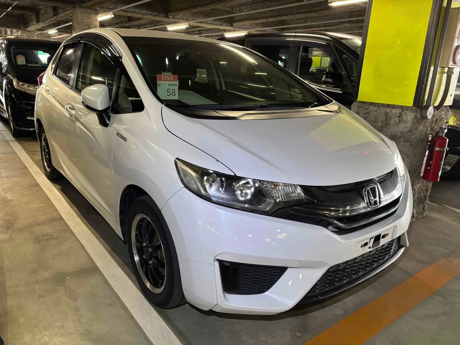 White Honda Fit Hybrid, front three-quarter view showing headlights, grille, Honda badge, and black aftermarket alloy wheels, parked in a covered auction lot