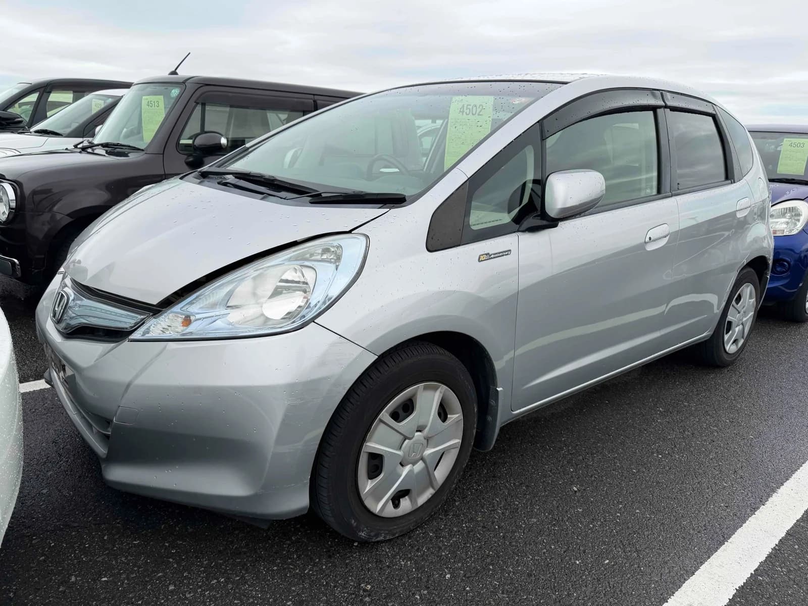 Silver Honda Fit Hybrid 10th Anniversary, front three-quarter view in the rain showing the Honda badge and alloy wheels, parked in an outdoor auction yard