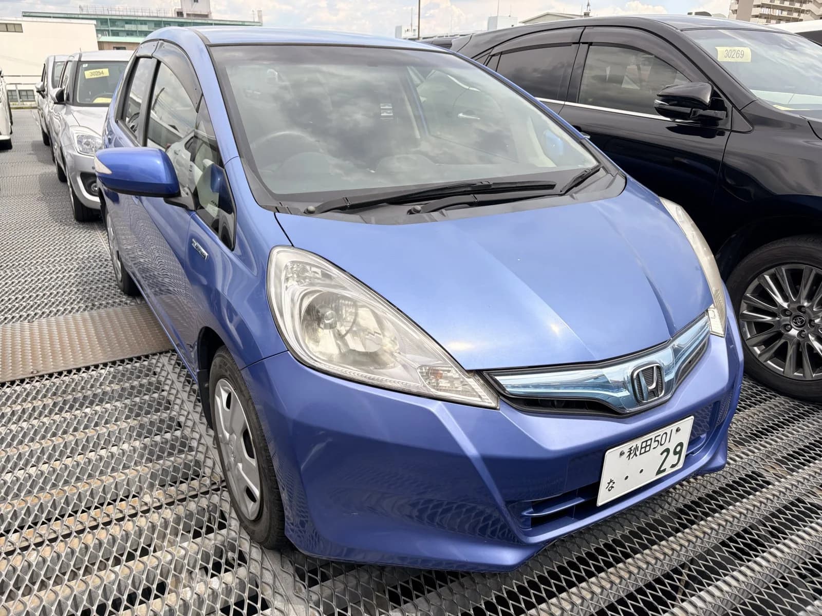 Blue Honda Fit Hybrid, front three-quarter view showing the Honda badge and chrome grille, parked among other cars in a gravel auction yard