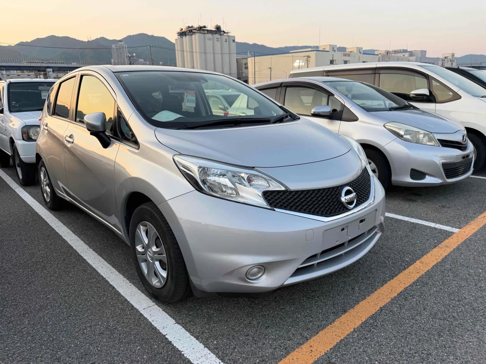 Silver Nissan Note, front three-quarter view at sunset showing the Nissan grille and headlights, parked among other cars in an outdoor auction yard
