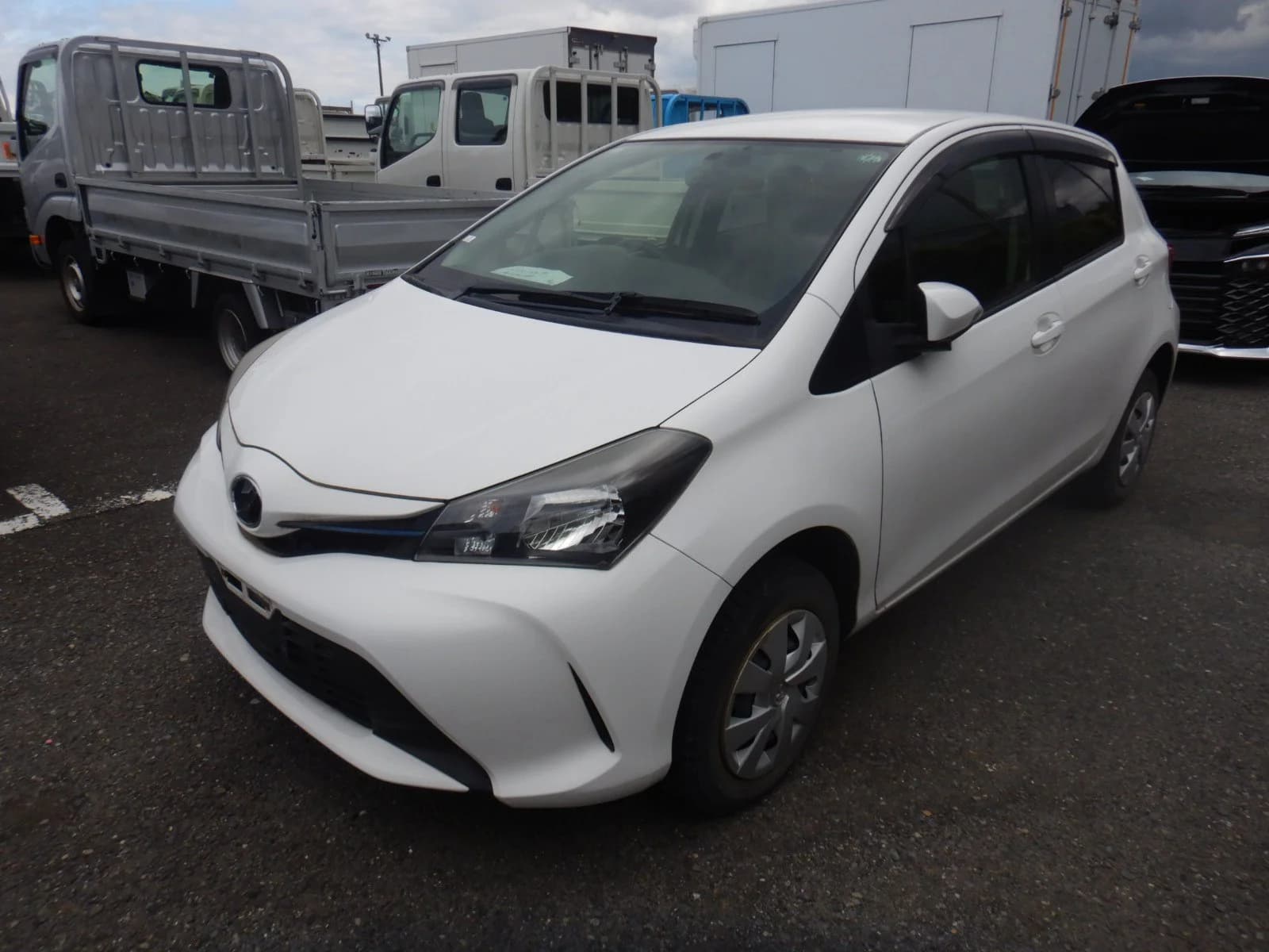 White Toyota Vitz hatchback, front three-quarter view showing headlights, grille, and Toyota badge, parked near delivery trucks