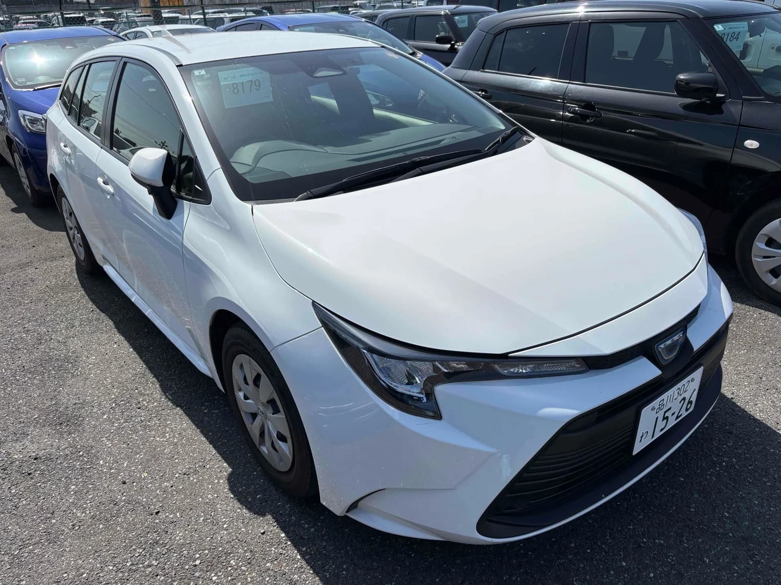 Toyota Corolla Touring hybrid sedan - Front-left 3/4 view showing white pearl paint, LED headlights, and modern design in rainy parking lot