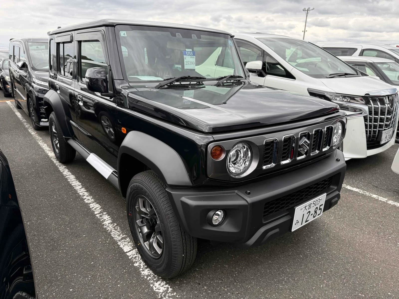 Black Suzuki Jimny Noble long-wheelbase 4x4, front three-quarter view showing chrome grille and round headlights, parked in an outdoor auction lot