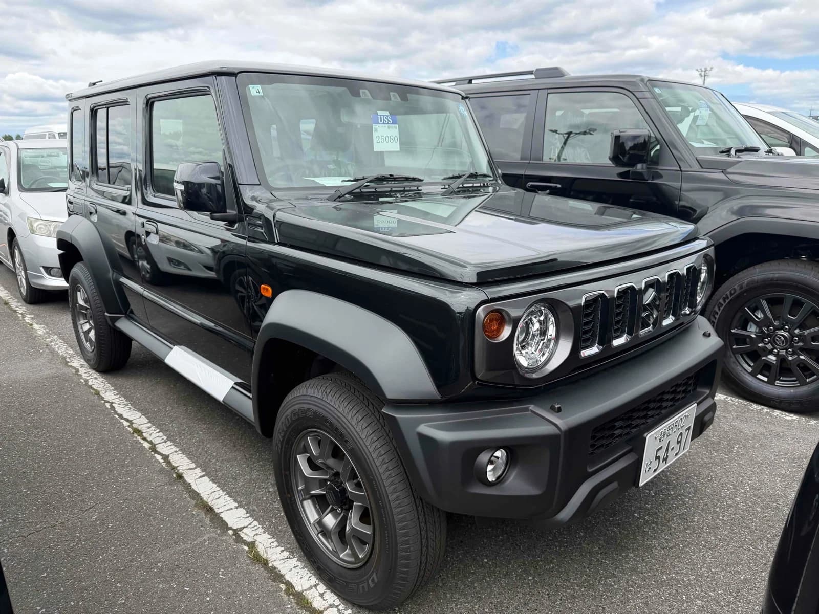 Black Suzuki Jimny Noble long-wheelbase 4x4, front three-quarter view showing chrome grille and round headlights, parked in an outdoor auction lot