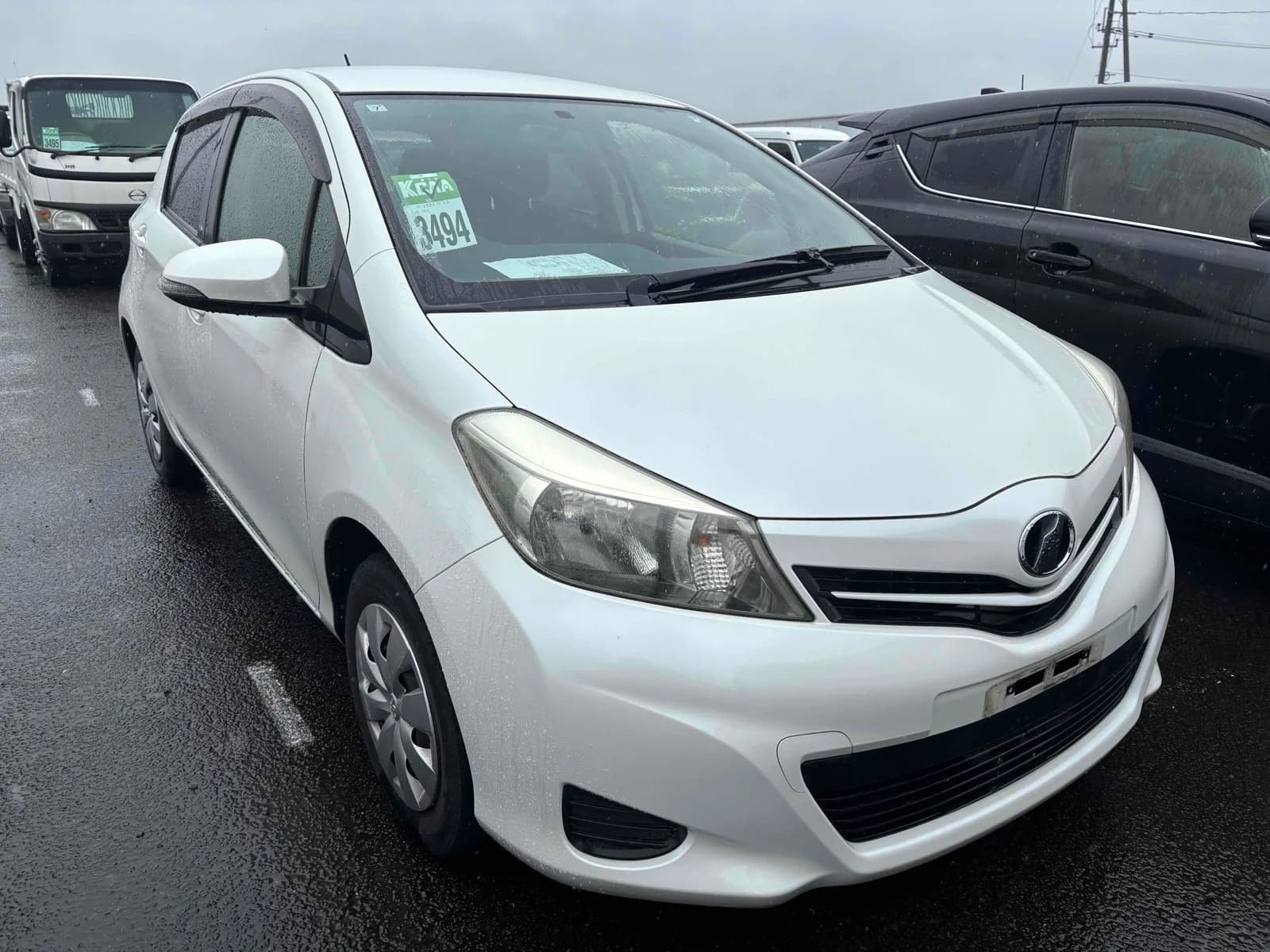 White Toyota Vitz hatchback, front three-quarter view showing headlights, grille, and Toyota badge, parked in an outdoor auction lot in the rain