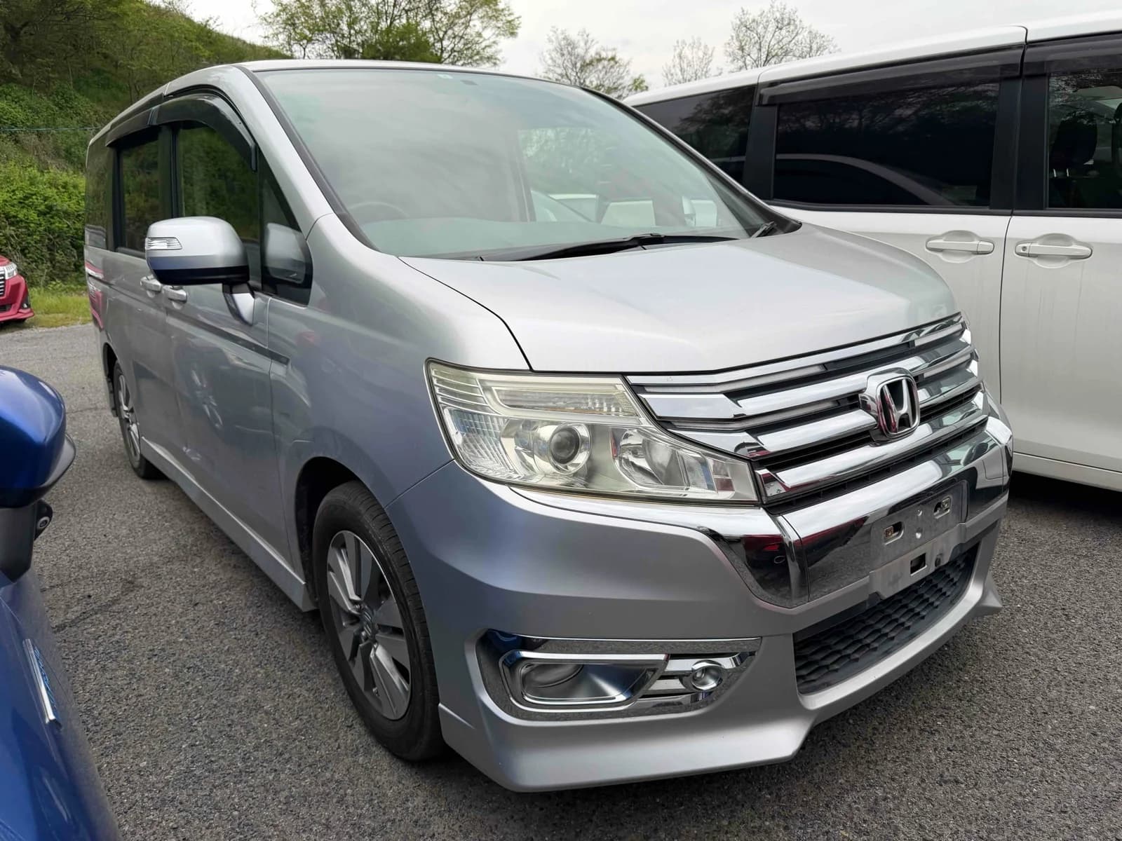 Silver Honda Stepwgn Spada minivan, front three-quarter view showing chrome grille, Honda badge, and headlights, parked among other minivans