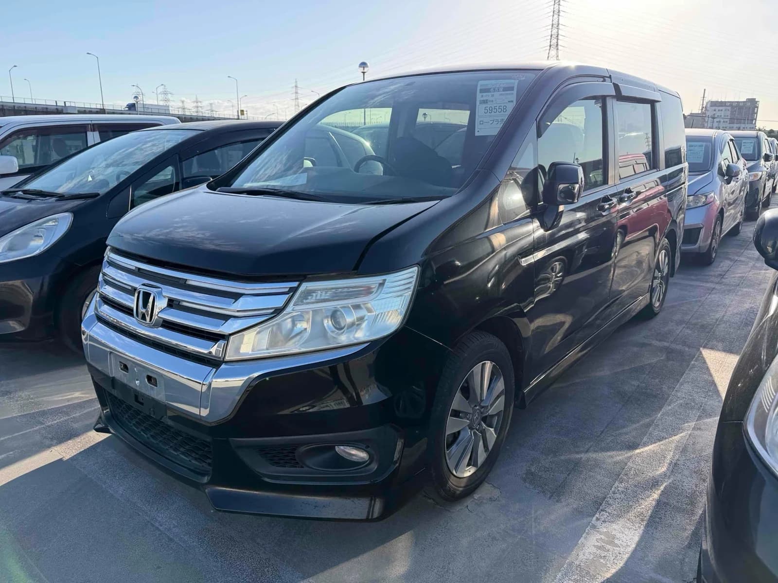 Black Honda Stepwgn Spada minivan, front three-quarter view showing chrome grille, Honda badge, and headlights, parked among other cars