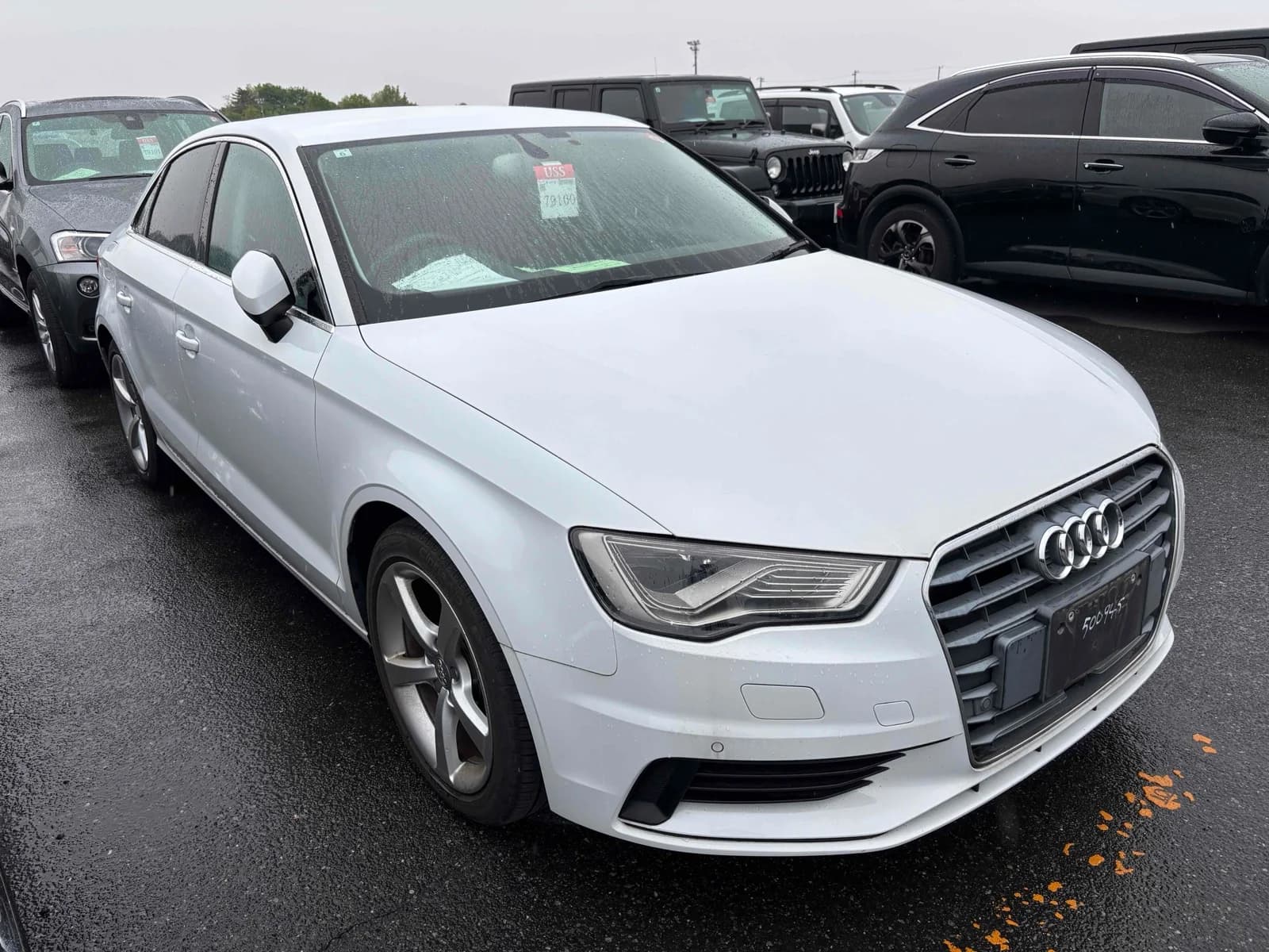 Front three-quarter view of a white Audi A3 sedan parked in an outdoor auction lot in the rain