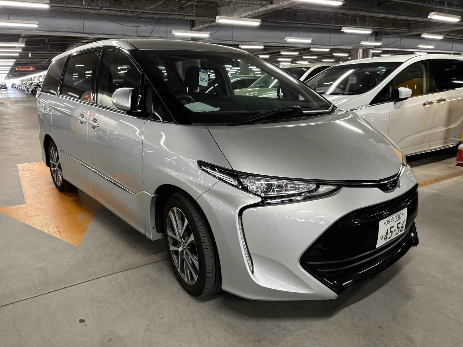 Silver Toyota Estima, front three-quarter view showing the swept-back headlights and black lower grille, parked in an indoor auction car park