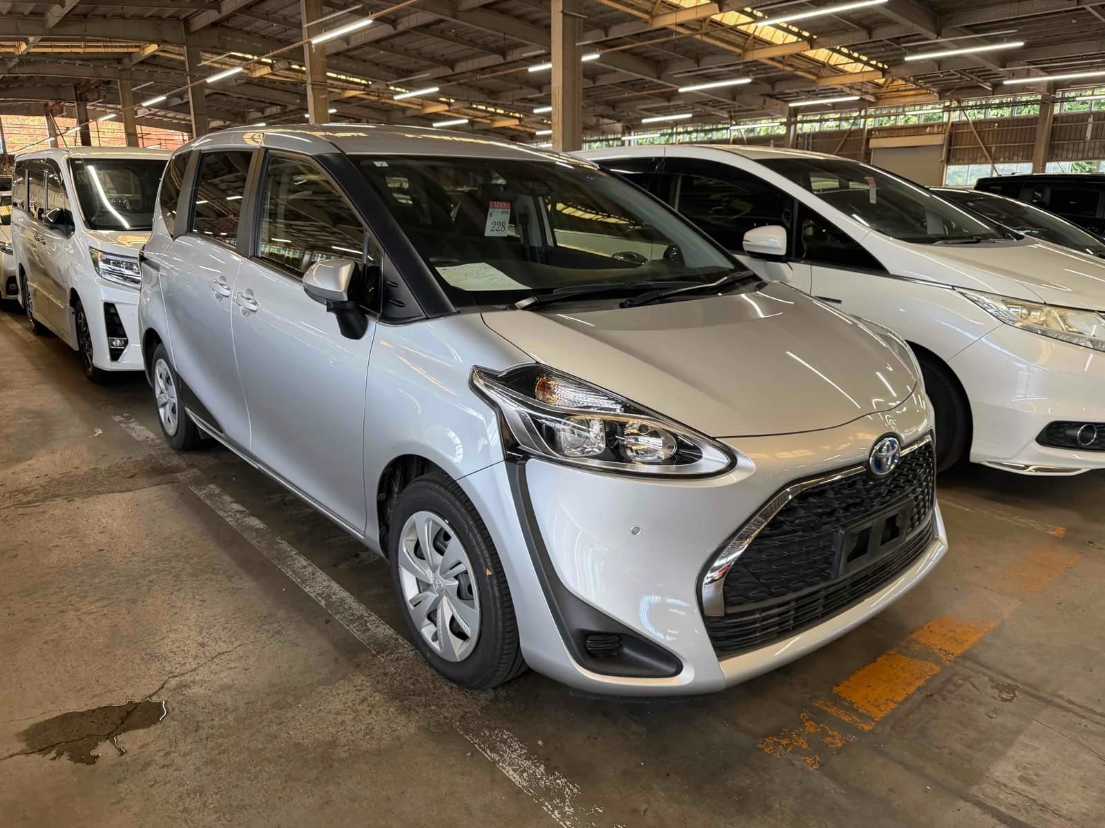 White Toyota Sienta compact minivan front-left three-quarter view in covered auction warehouse with industrial metal roof structure