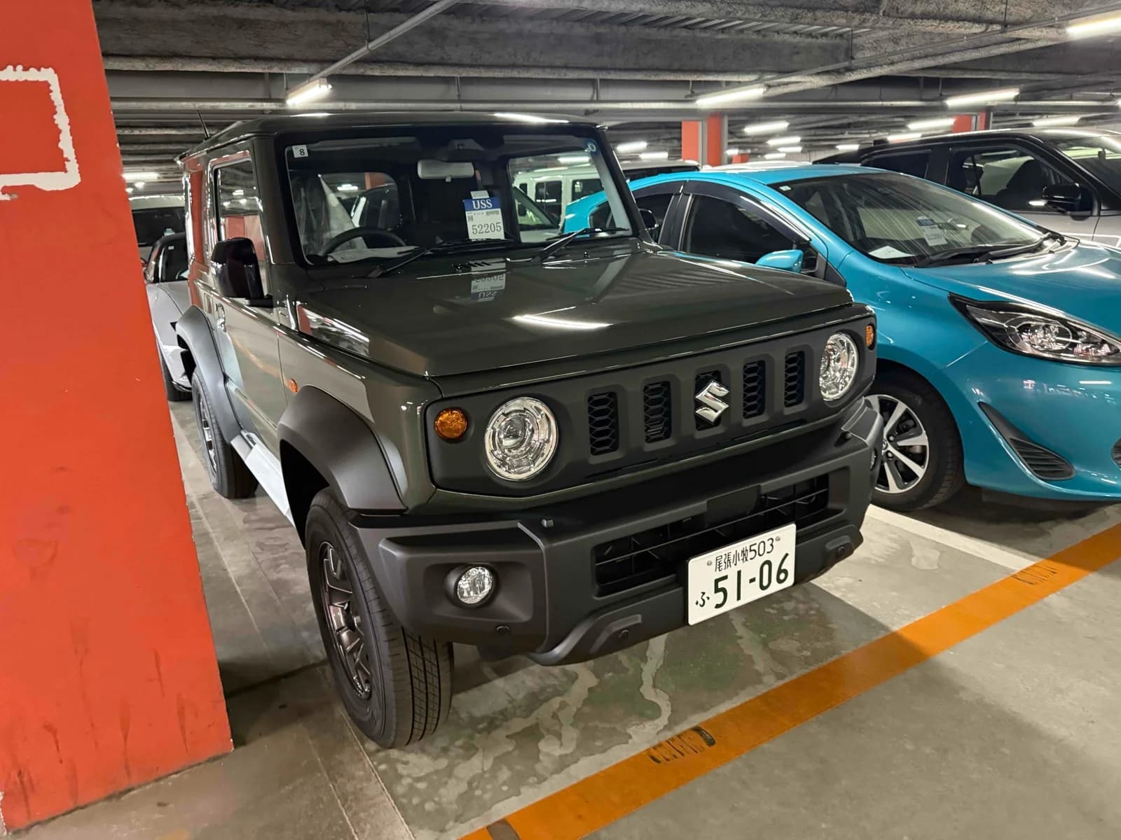 Olive green Suzuki Jimny compact SUV - front-left 3/4 view in parking garage showing the round LED headlights and distinctive vertical grille