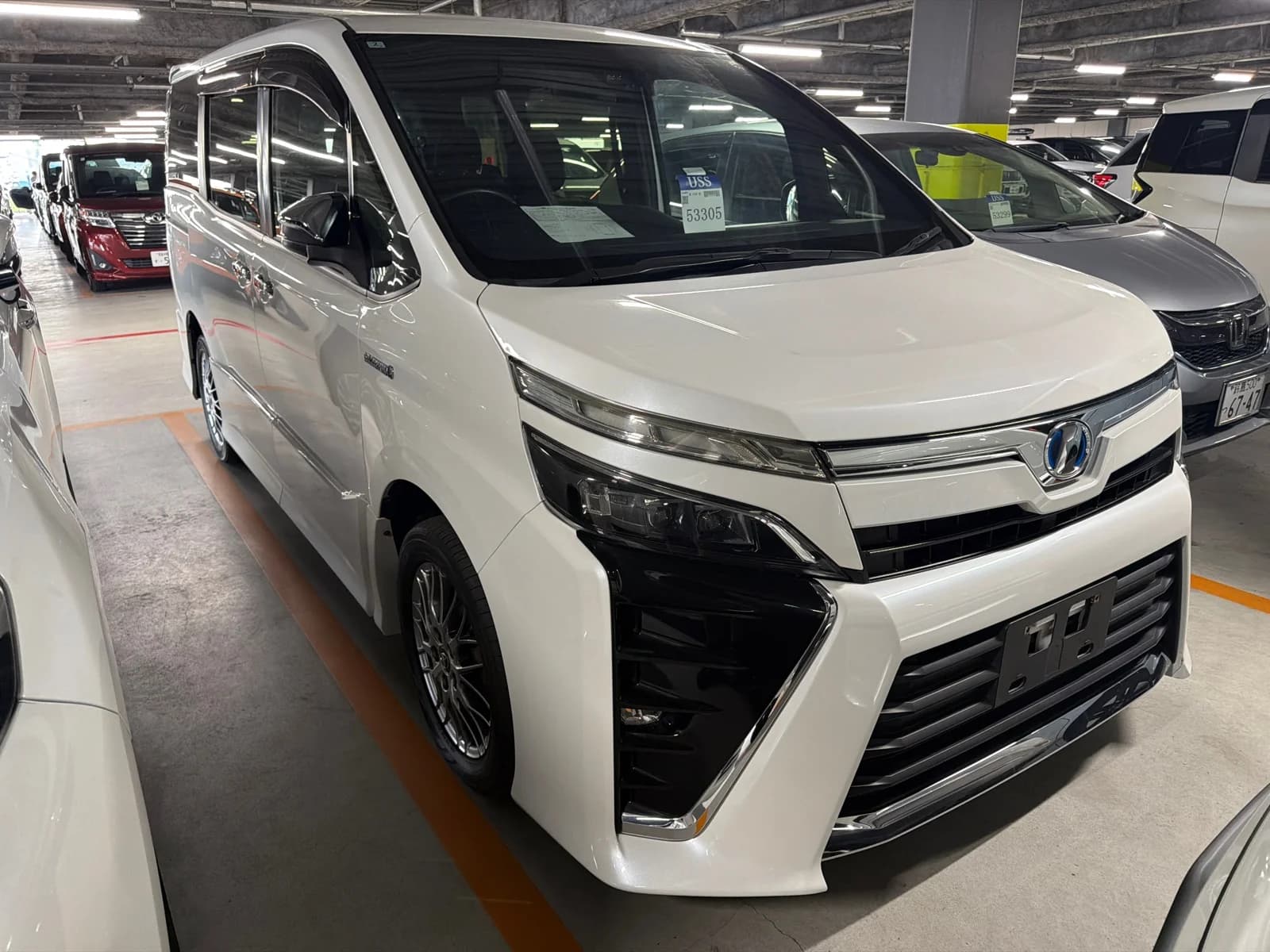 2018 Toyota Voxy Hybrid ZS Kirameki in pearl white - front-right 3/4 view with LED headlights, chrome-trimmed grille, blue hybrid Toyota emblem, window visors, and aftermarket mesh alloy wheels