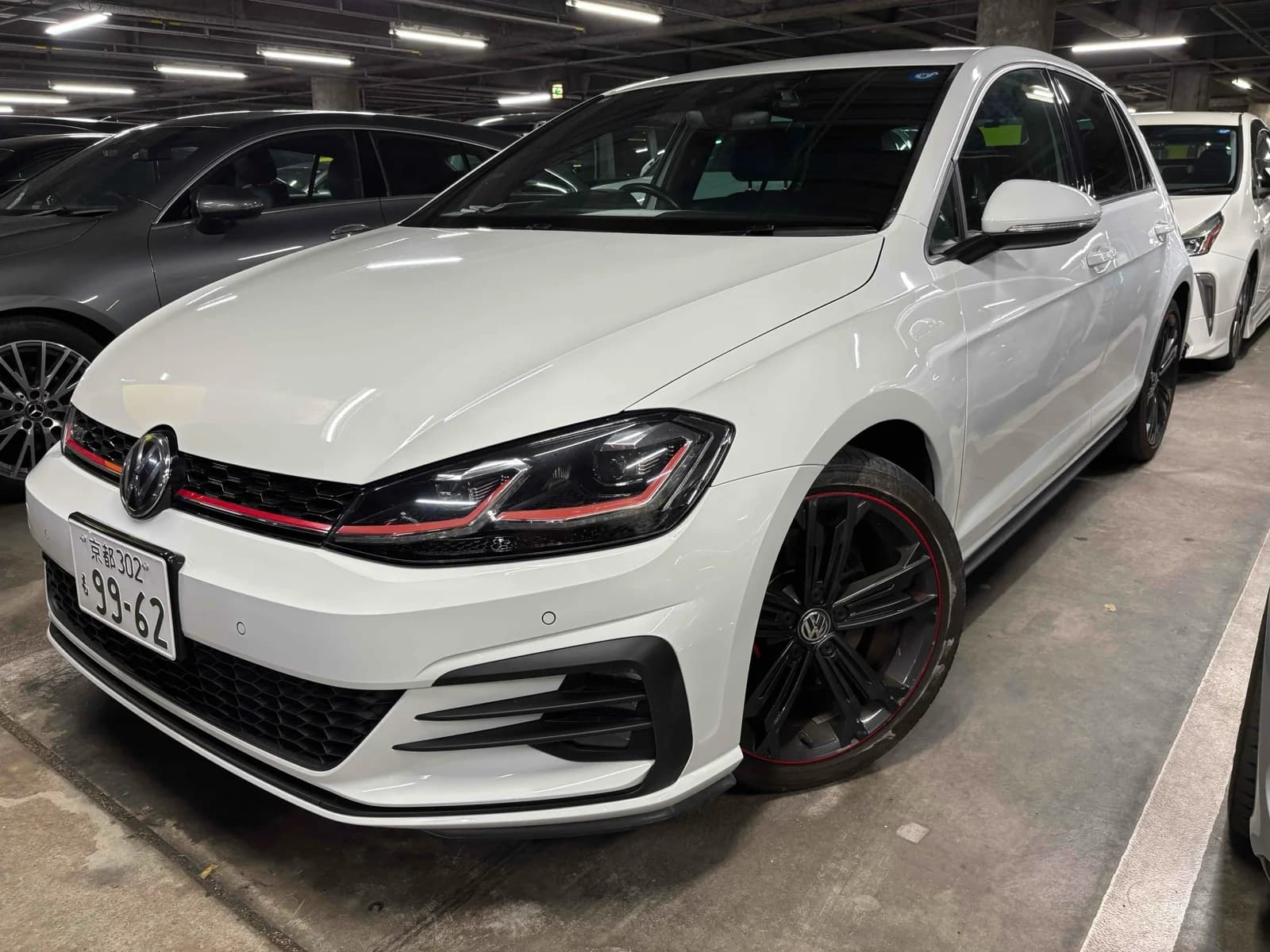 White Volkswagen Golf GTI hatchback front-right three-quarter view in indoor parking garage with red grille stripe and black alloy wheels