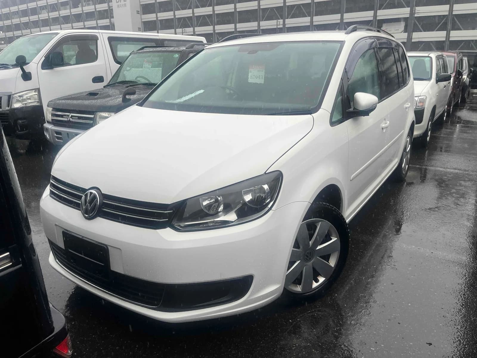 White Volkswagen Touran minivan front-right three-quarter view at outdoor auction lot in the rain with roof rails