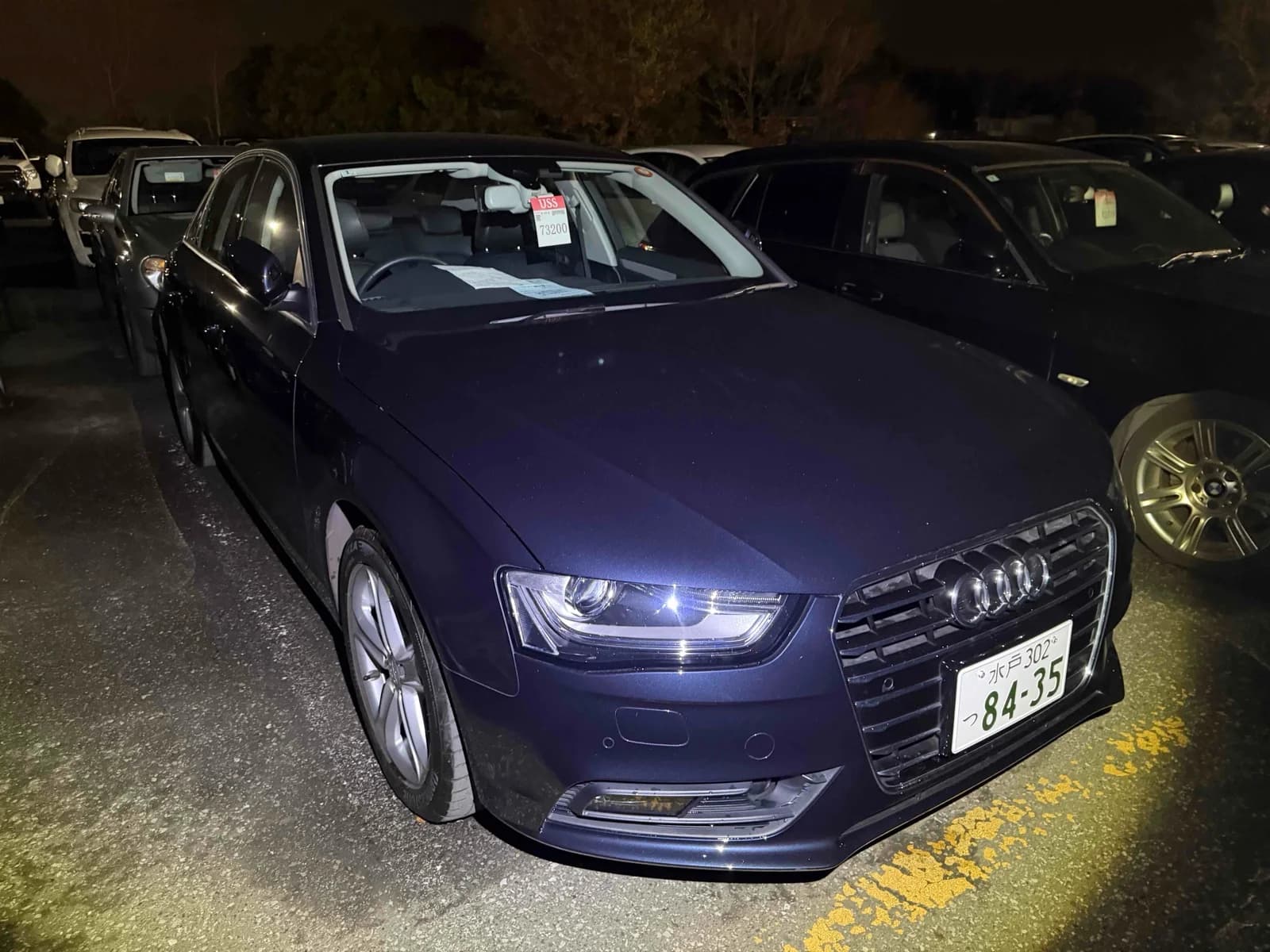 Dark blue Audi A4 sedan front-right three-quarter night view at auction yard with USS price tag on windshield