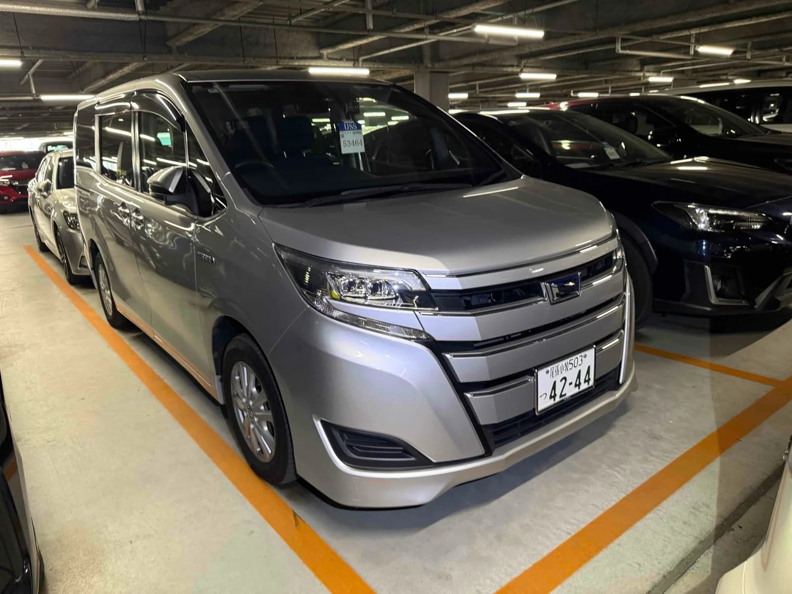 Silver Toyota Noah hybrid minivan front-right three-quarter view in indoor parking garage with chrome grille