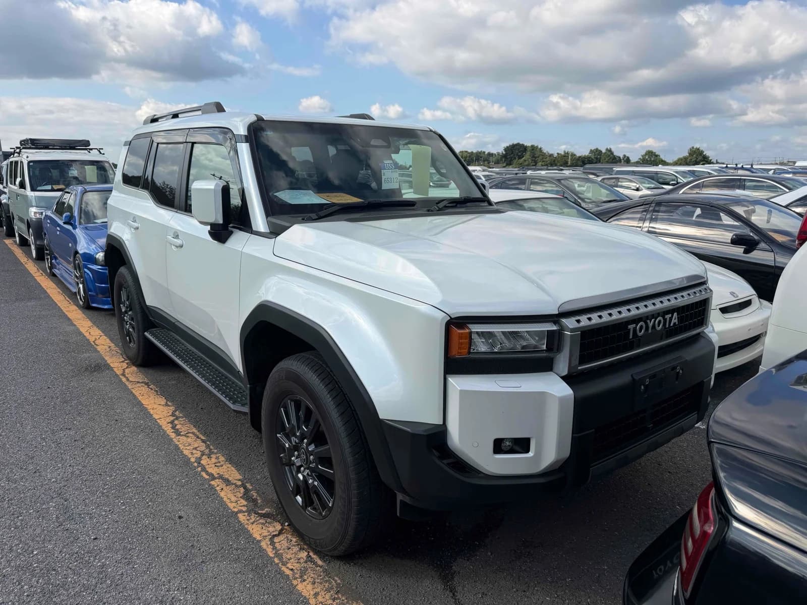 Toyota Land Cruiser 250 SUV - Front-right exterior view in a daytime auction lot with white paint, black roof rails, LED headlights, and black steel wheels