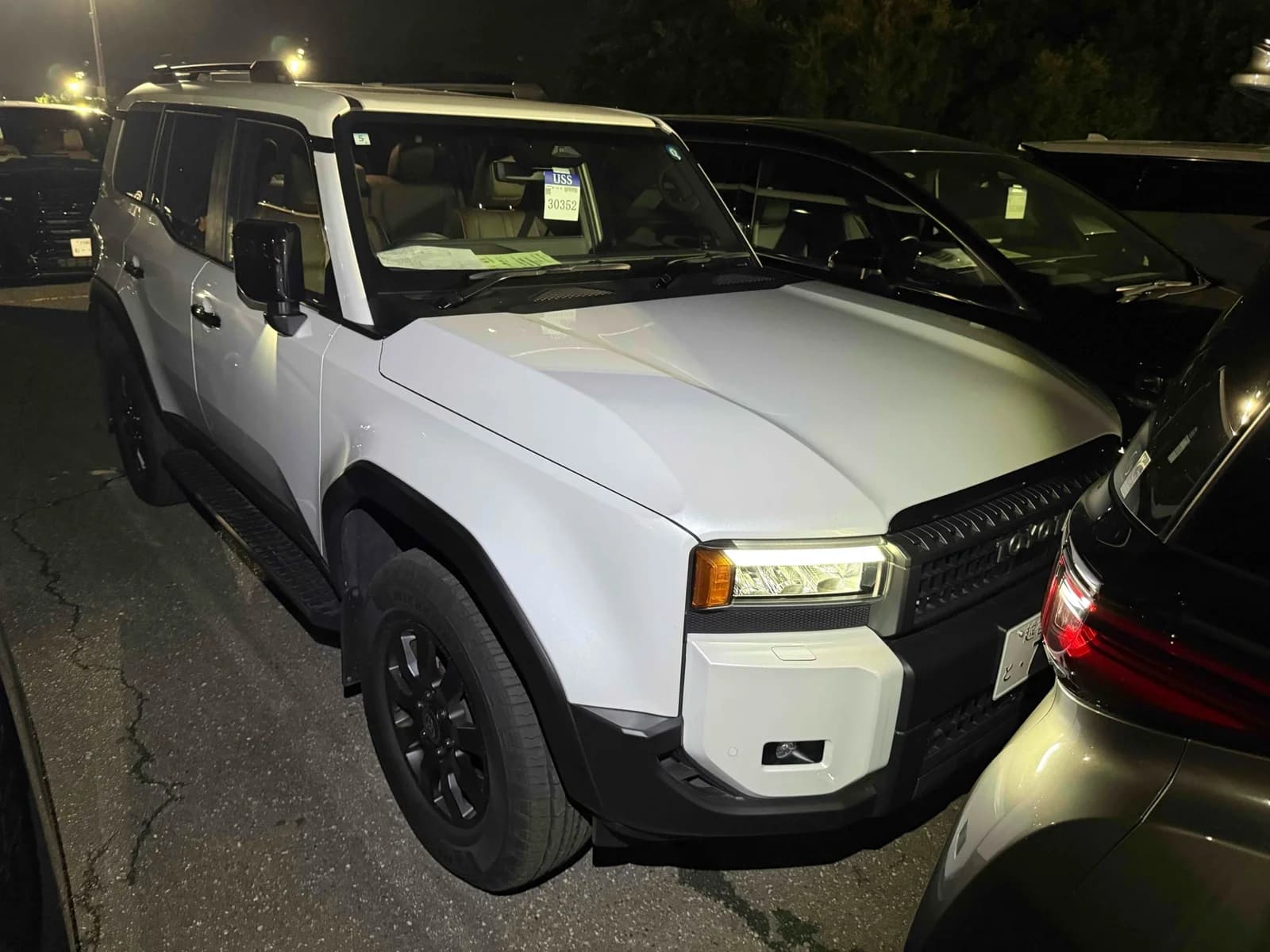 Toyota Land Cruiser 250 SUV - Front-left exterior view at night in white paint with matte black hood, LED headlights, and black steel wheels