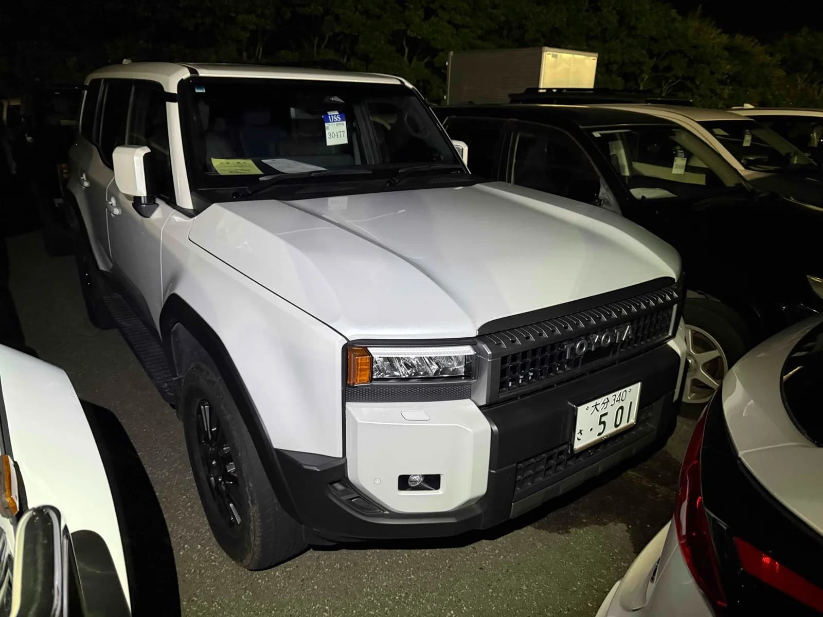 Toyota Land Cruiser 250 SUV - Front-left exterior view at night in white paint with matte black hood, LED headlights, and black steel wheels