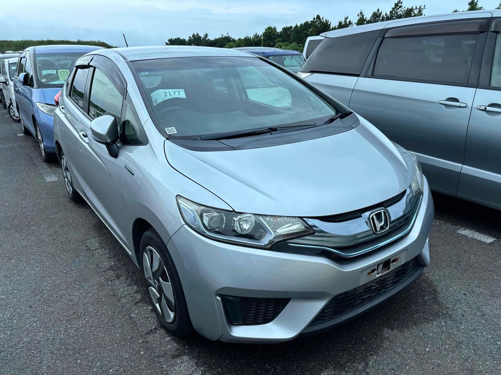 Honda Fit Hybrid hatchback - Front-right exterior view in an auction lot with silver paint, black window visors, and alloy wheels