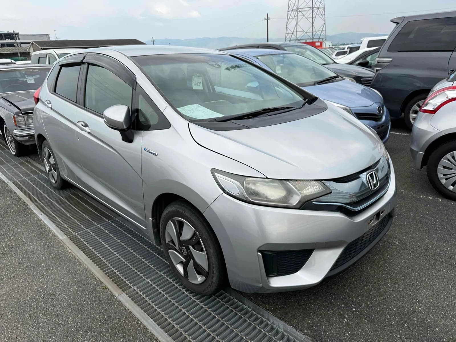 Honda Fit Hybrid hatchback - Front-right exterior view in an auction lot with silver paint, black window visors, and alloy wheels