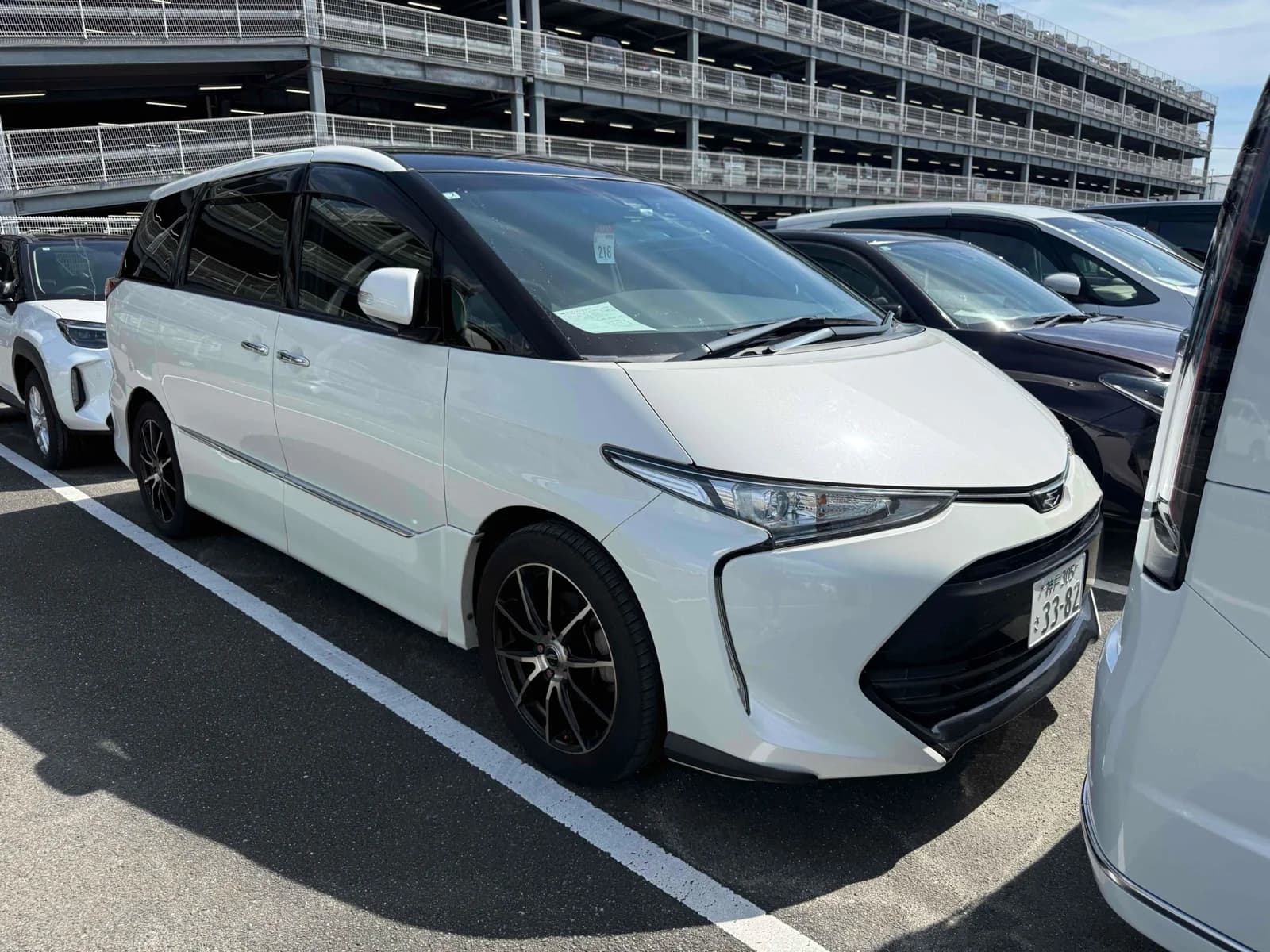 Toyota Estima minivan - Front-right exterior view in a parking lot with pearl white paint, black roof rails, and dark alloy wheels with red lug nuts