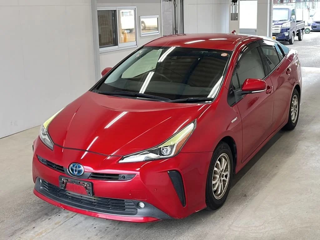 Toyota Prius hatchback - Front-left exterior view with red metallic paint, LED headlights, and chrome grille in an indoor showroom