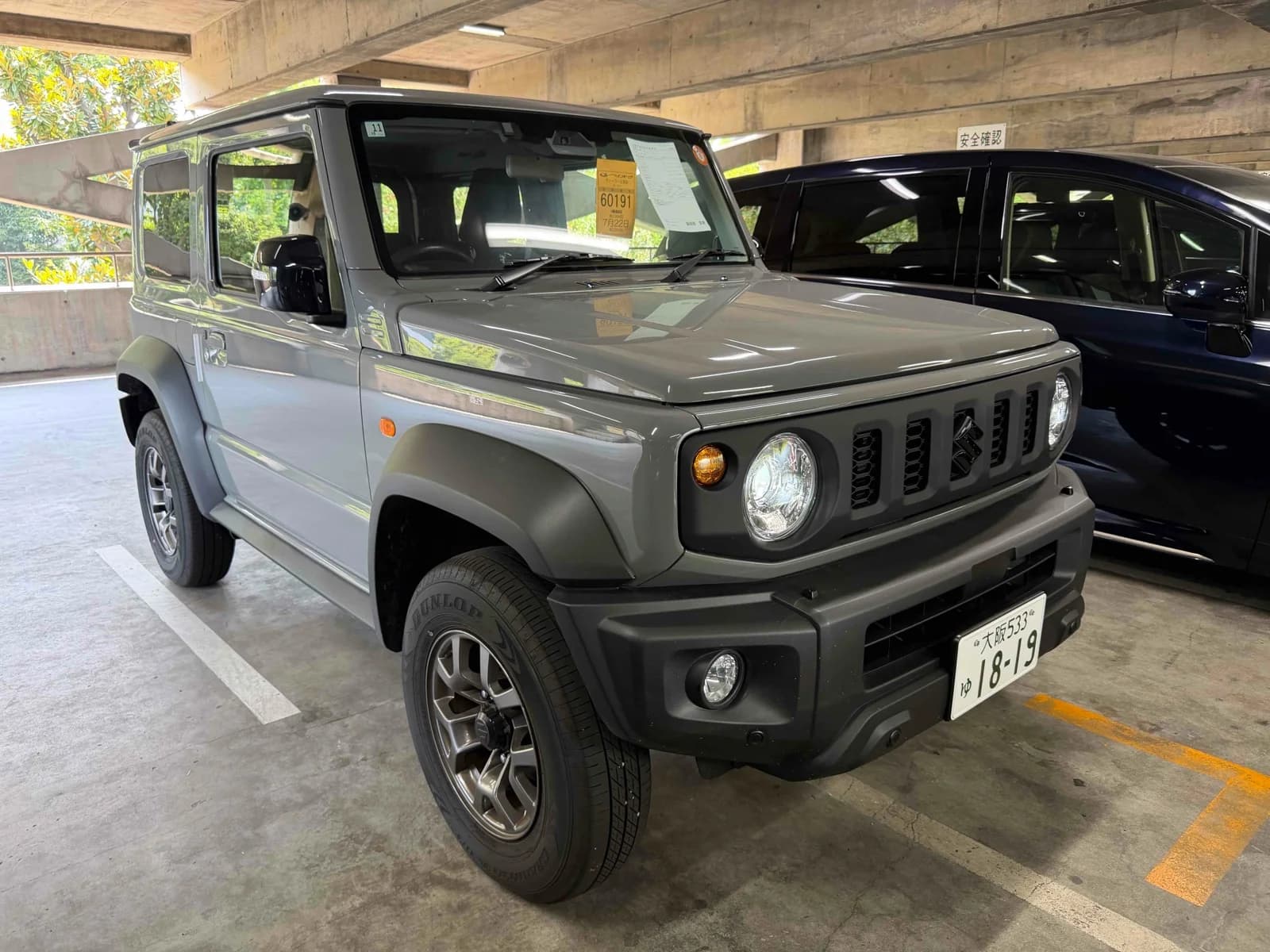 Suzuki Jimny Sierra JB74W - Front-right three-quarter view in gray with round LED headlights, black slotted grille, fender flares and Dunlop tires on bronze alloy wheels