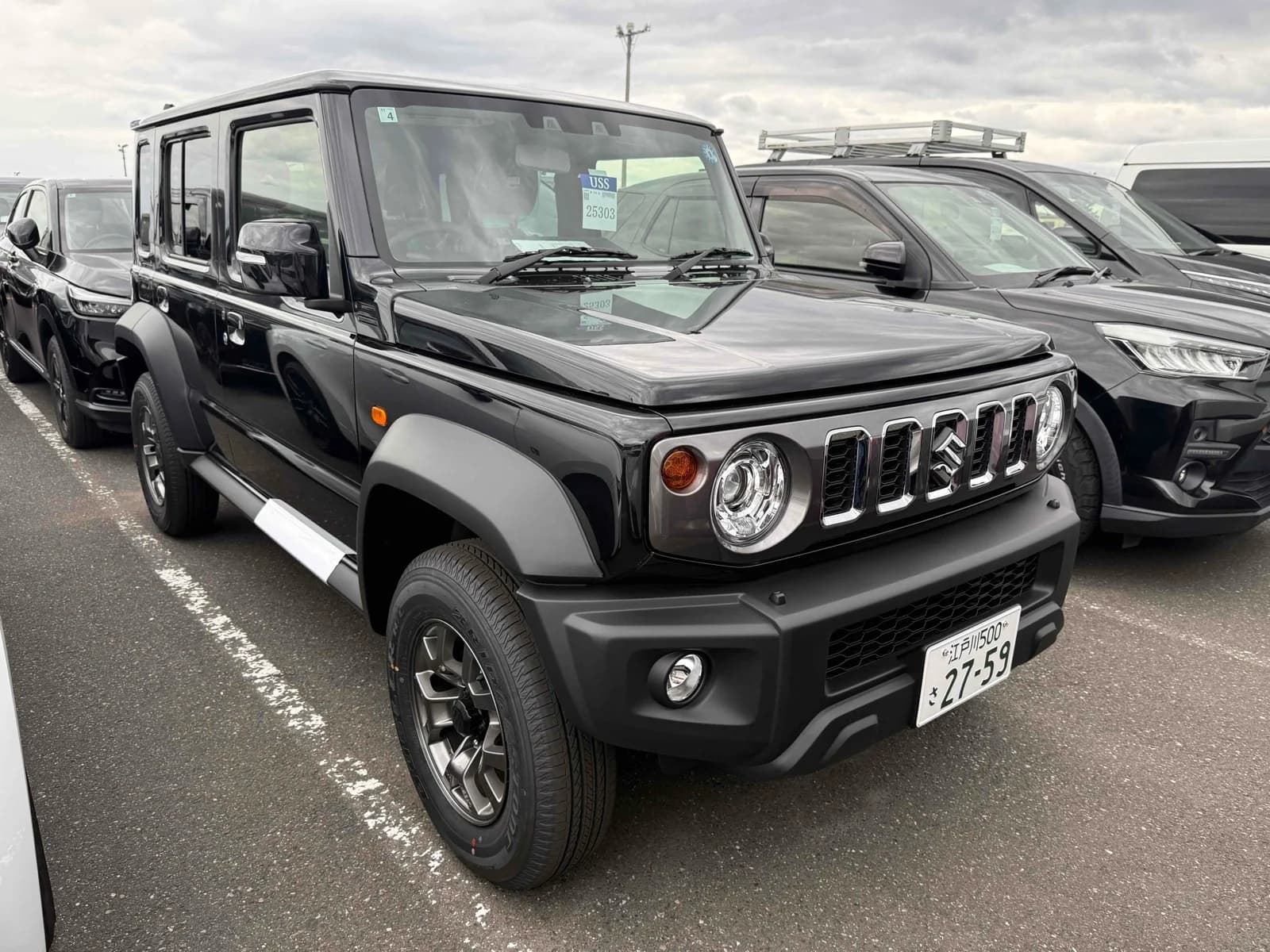 2026 Suzuki Jimny Nomade 5-door 4WD in black - front-right 3/4 view with chrome-slotted grille, round LED headlights, fender flares, and gunmetal alloy wheels
