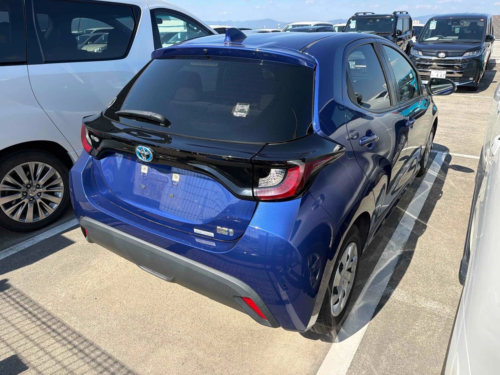 2021 Toyota Yaris Hybrid X in metallic blue - rear-right 3/4 view with gloss black tailgate trim, LED tail lights, blue hybrid Toyota badge, and steel wheels with covers